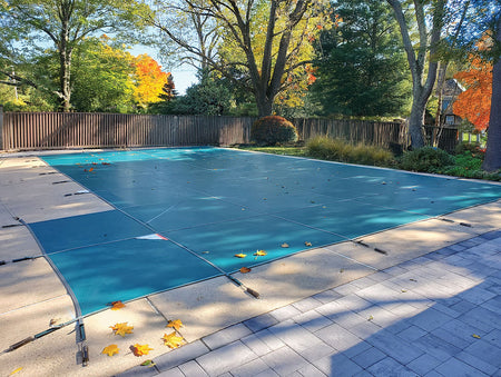 Pool with a blue cover in a backyard setting with trees and a fence.